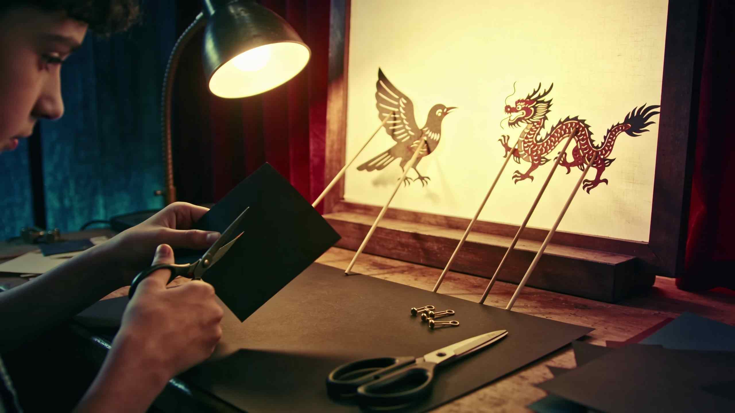 Children cutting shadow puppets from black card at a workshop table, a bird and a dragon puppet on rods in front of a lit screen