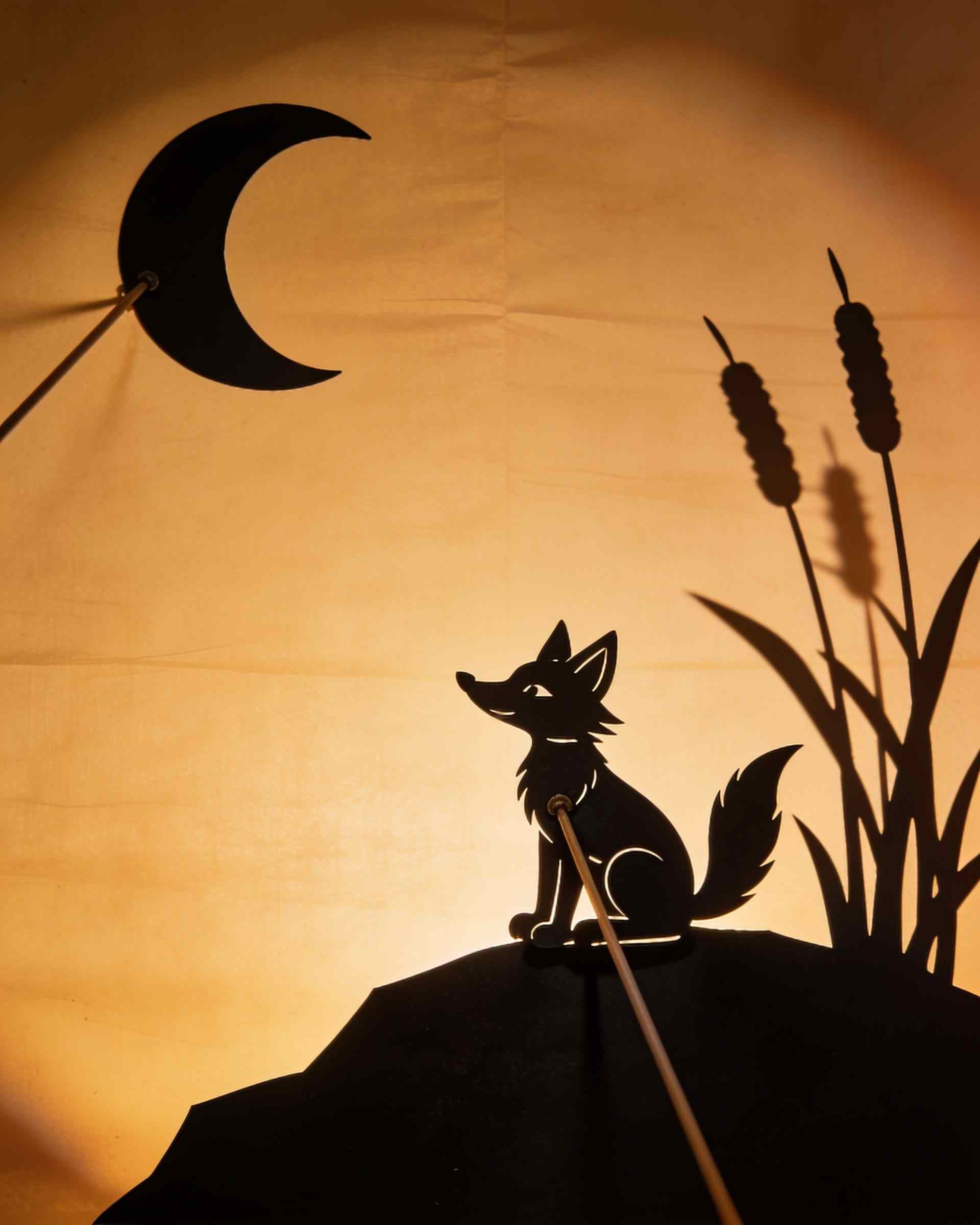 A shadow scene on a glowing screen: a fox puppet sitting on a hill under a crescent moon, reeds beside it